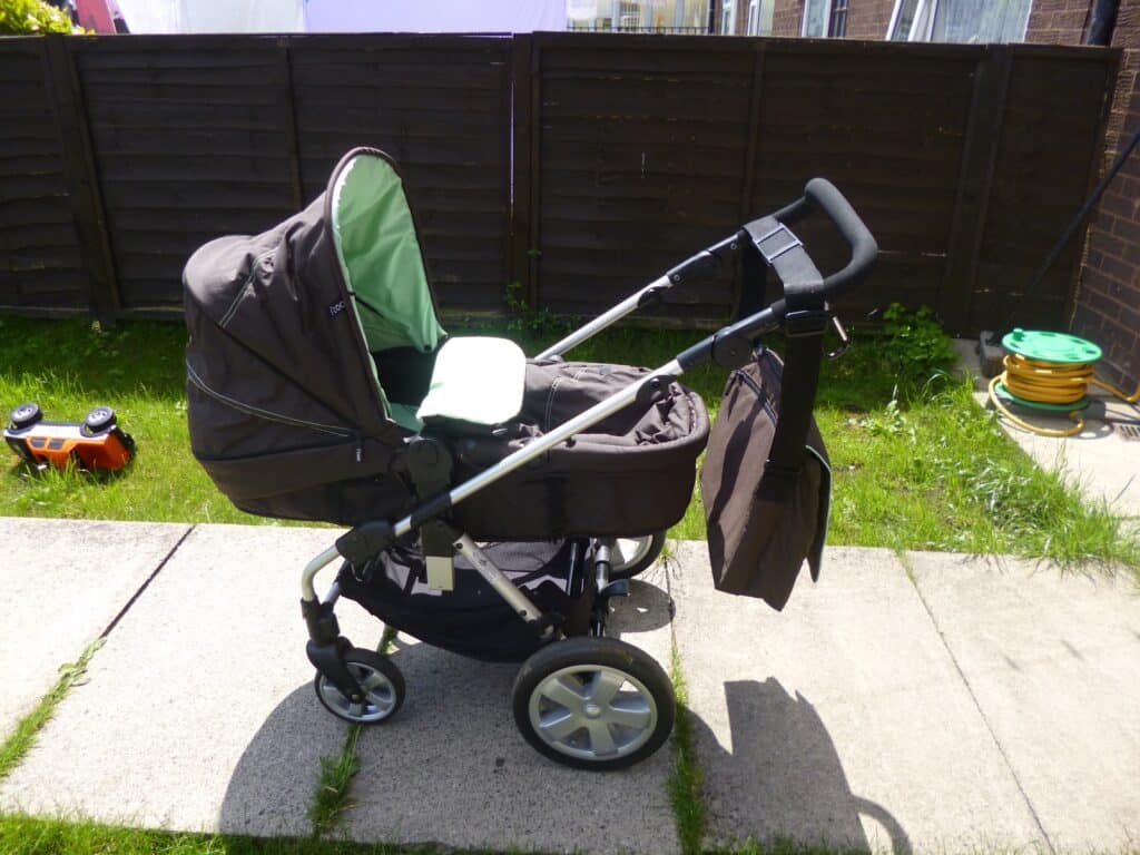 A brown I'coo Pacific pram with a green interior is parked on a concrete path in a grassy garden. A matching bag hangs from the handle. Nearby, there is a yellow garden hose and a small orange toy vehicle.