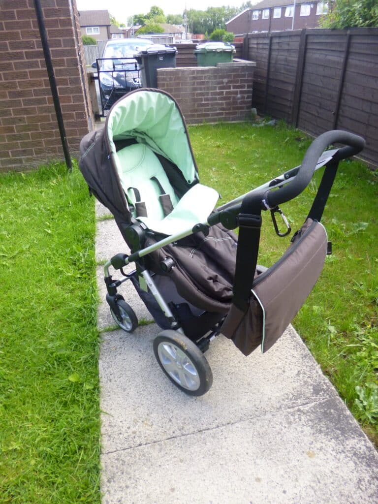 A green and brown Hauck 2 in 1 pram with a matching bag attached to the handle is positioned on a narrow concrete path between grassy lawns and brick and wooden fences in a residential area.