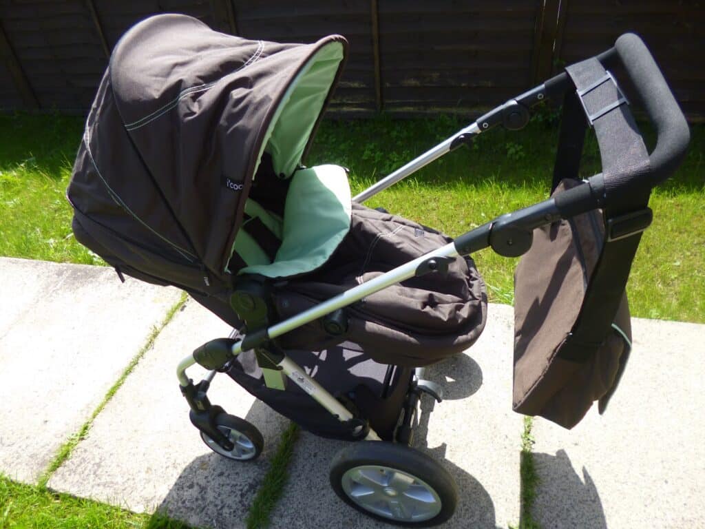 A brown and mint green Hauck 2 in 1 pushchair with a sunshade is parked on a paved path next to green grass. A matching bag hangs from the handle, and the pram is empty.