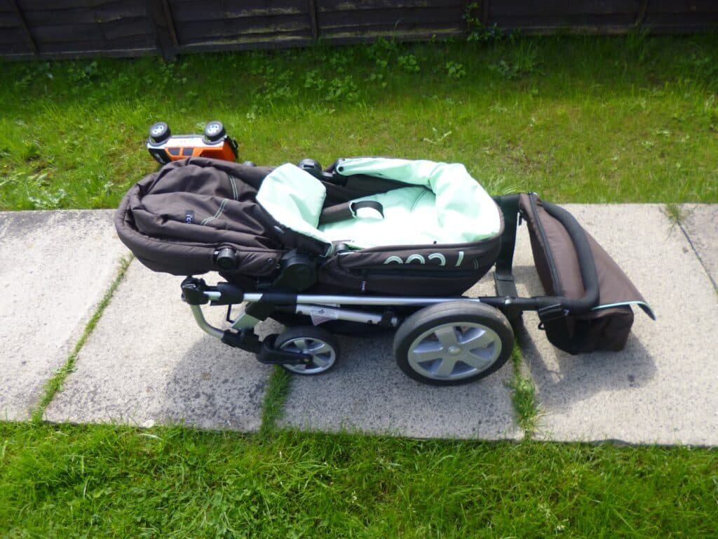 A black and mint green Hauck 2 in 1 pushchair is parked on a paved walkway, with grass on either side and a brown fence in the background. A small toy car is visible behind the pushchair.