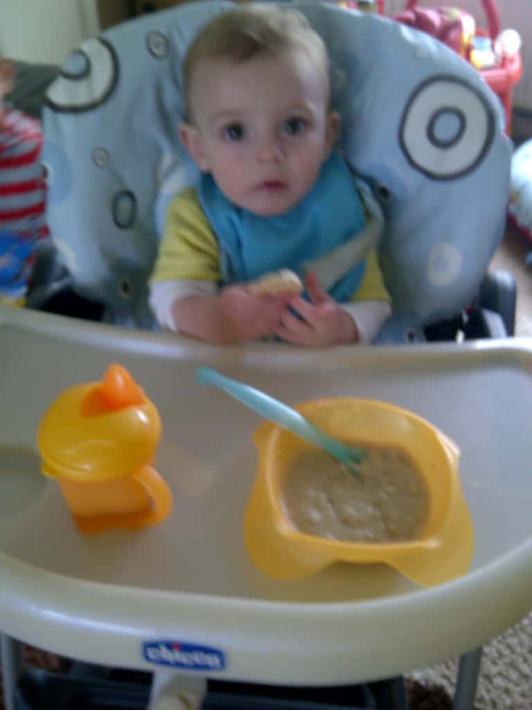 A baby sits in a highchair wearing a blue bib, holding food. On the tray are Vital Basics: a yellow bowl with a blue spoon and a yellow beaker. The baby looks up towards the camera—Simply Everything You Need for mealtime.