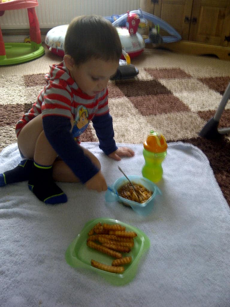 A young child sits on a white towel indoors, eating food from a bowl with a fork. Surrounded by Simply Everything You Need—crinkle-cut chips, a green beaker, and toys—the cosy room features carpeted comfort for playful moments.