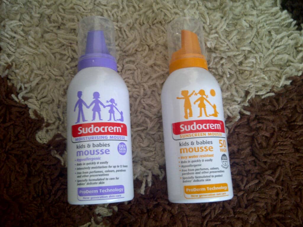 Two bottles of Sudocrem Mousse for kids and babies on a brown and beige carpet: one is a moisturising mousse with a purple cap, and the other is a suncream mousse SPF 50 with an orange cap—perfect skin care essentials.