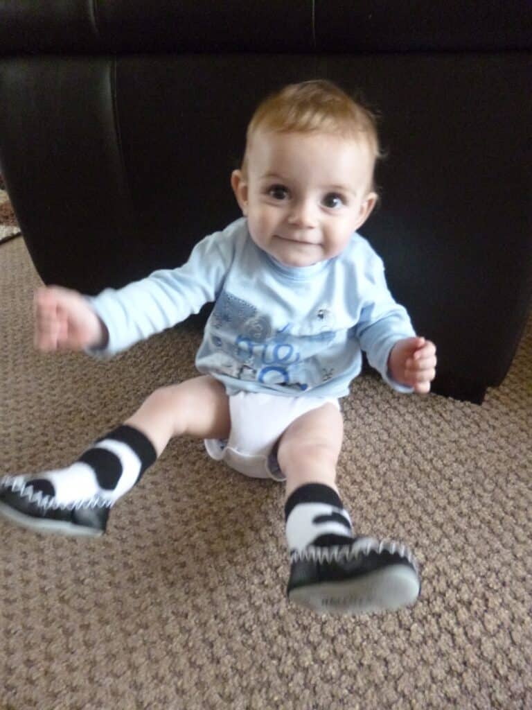 A smiling baby with light brown hair sits on a beige carpet, wearing a light blue shirt, white nappy, and black-and-white socks, with arms raised and a dark sofa behind them.