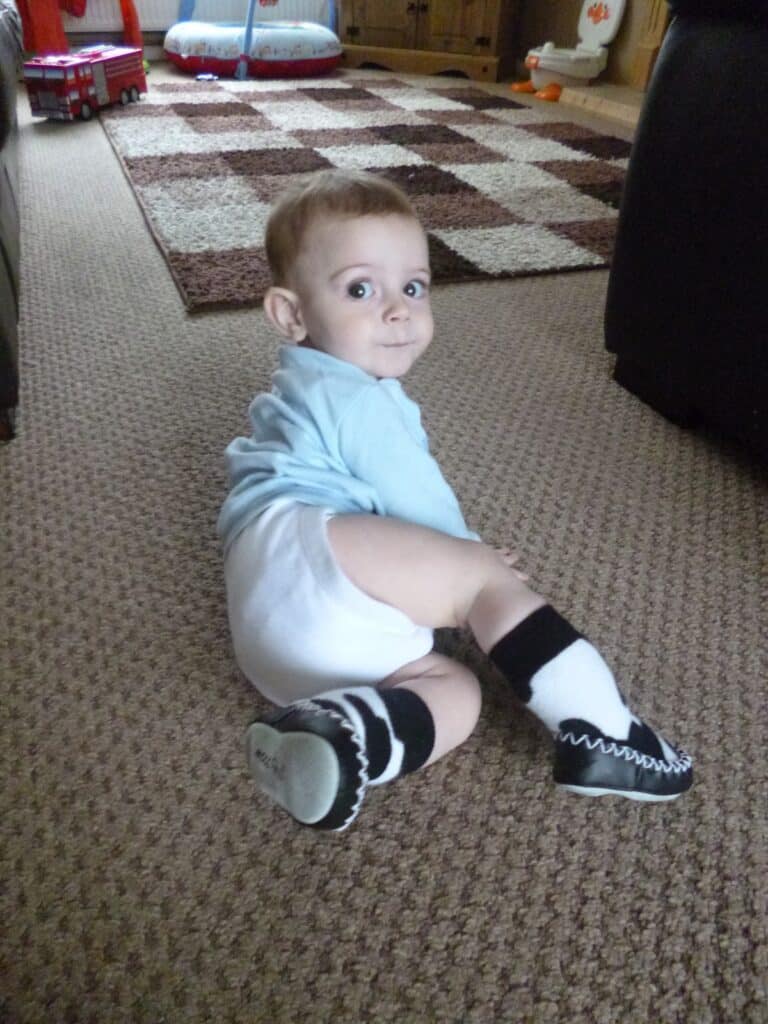 A baby in a light blue shirt, white nappy, and black-and-white socks sits on a carpeted floor, looking back over their shoulder in a cosy living room.