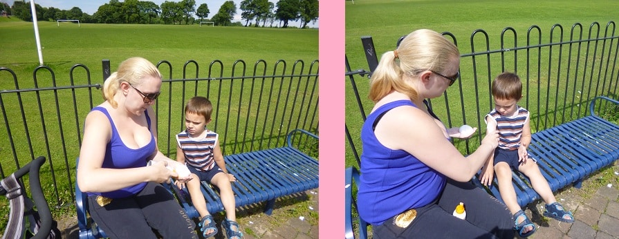 A woman applies suncream and cares for a young boy’s skin as he sits on a blue bench in a sunny park. The boy looks down while she gently puts cream on his face, showing her dedication to proper skin care. A pushchair and open field are in the background.