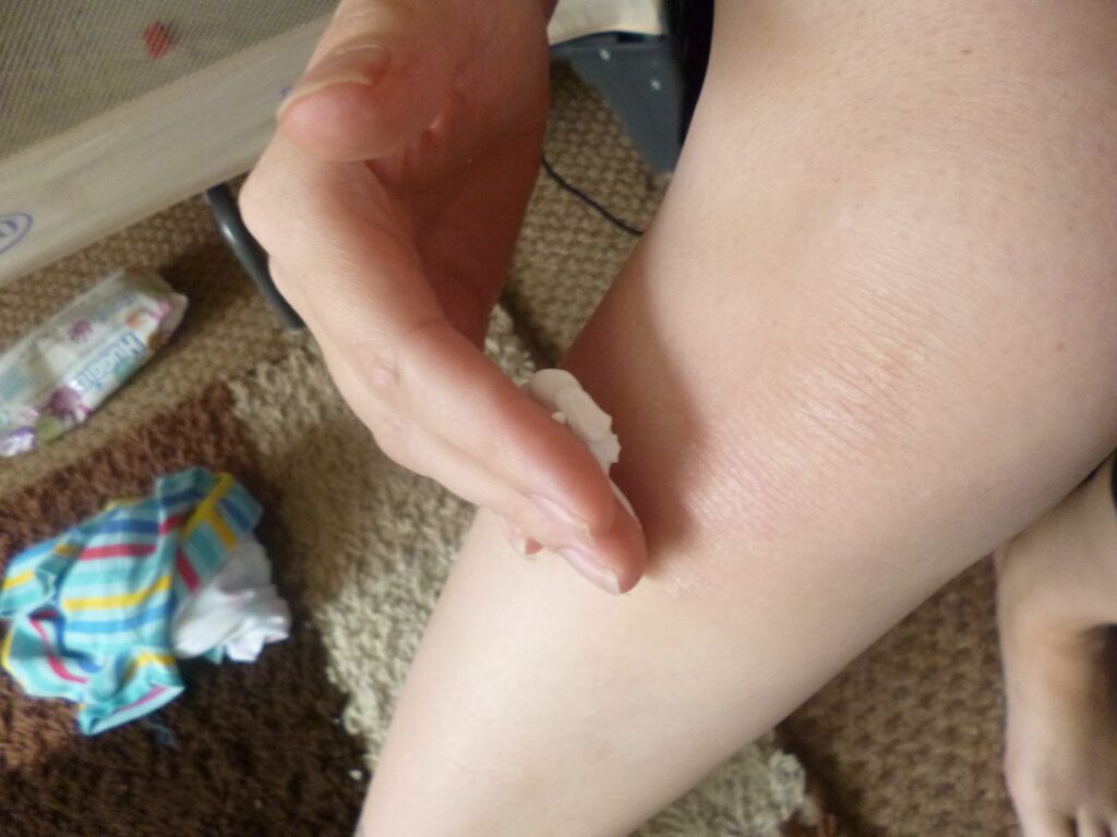A person is applying Sudocrem Mousse to their leg with their fingers as part of their skincare routine. A colourful striped cloth and some objects are on a brown textured carpet in the background.