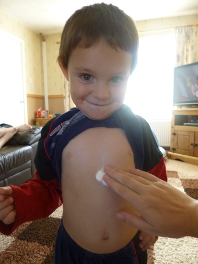A young boy lifts his shirt and smiles at the camera whilst someone applies a dab of Sudocrem Mousse to the centre of his chest in a living room setting.