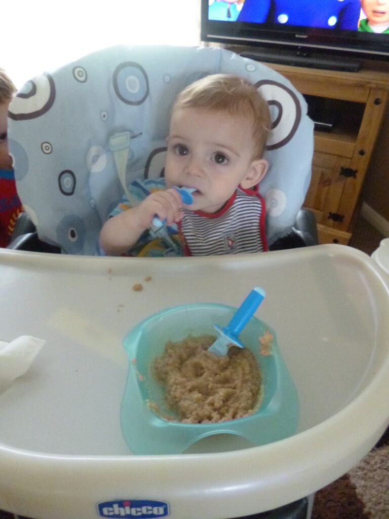 A baby sitting in a highchair holds a blue spoon from the MAM Dipper Set and looks at the camera, with a bowl of baby food and another blue spoon on the tray in front of them.