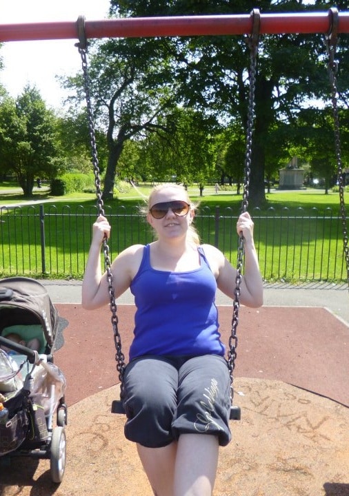 A woman wearing sunglasses and a blue vest sits on a swing in a sunny park, enjoying some post-pregnancy relaxation. Beside her is a pushchair, with lush green trees in the background.