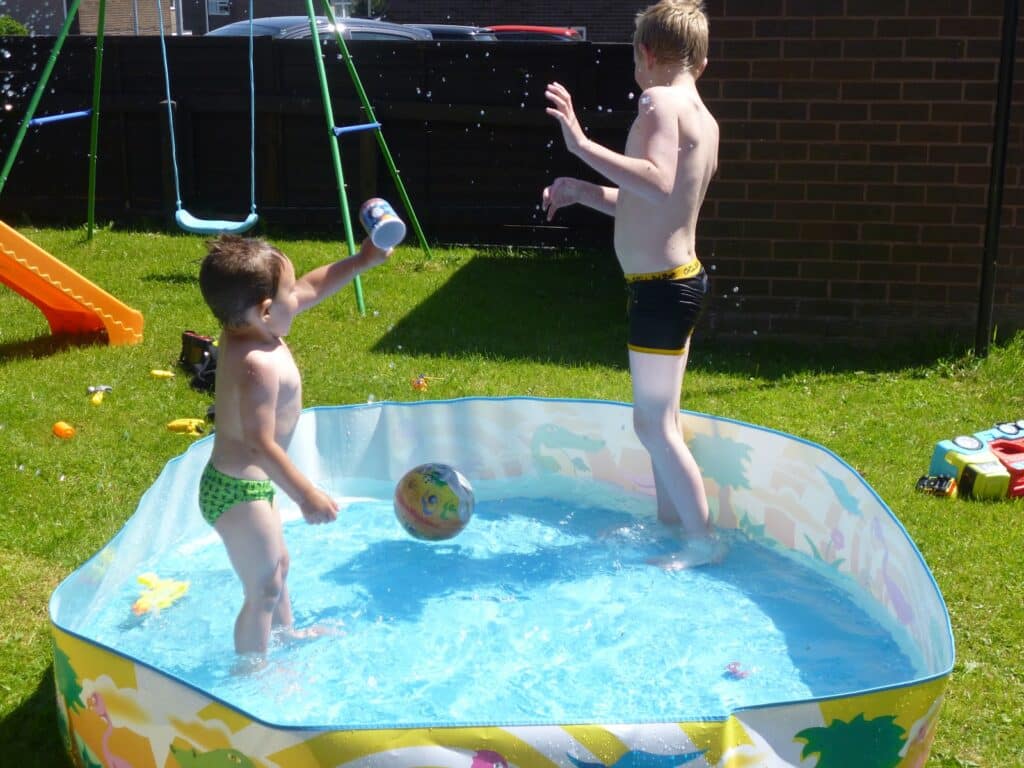 Two children play in a small back garden paddling pool on a sunny Family Day. One boy splashes water with a cup while the other stands nearby. Toys and a swing set are visible in the grassy garden, creating a fun scene of family activities.