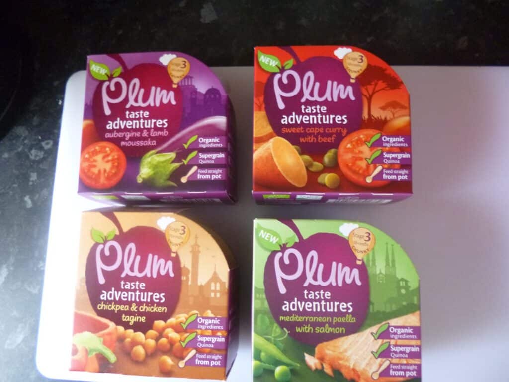 Four Plum Taste Adventures ready-meal pots are arranged on a worktop, featuring flavours: aubergine & lamb moussaka, sweet cape curry with beef, chickpea & chicken tagine, and Mediterranean paella with salmon.