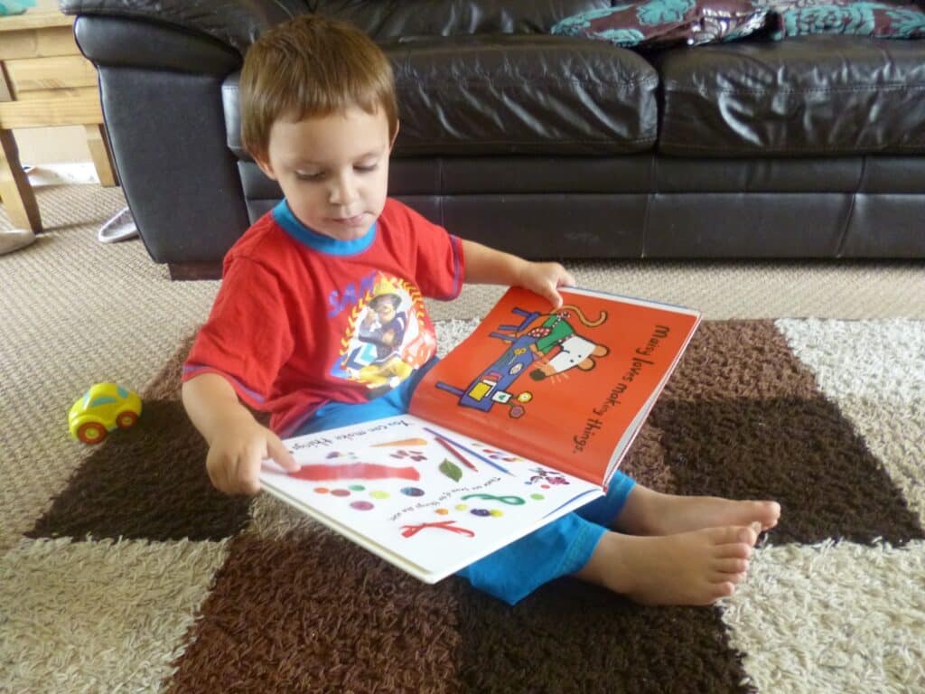 A young child sits barefoot on a brown and cream chequered rug, reading a colourful Maisy craft book. A small toy car is nearby, and a dark brown leather sofa is in the background.