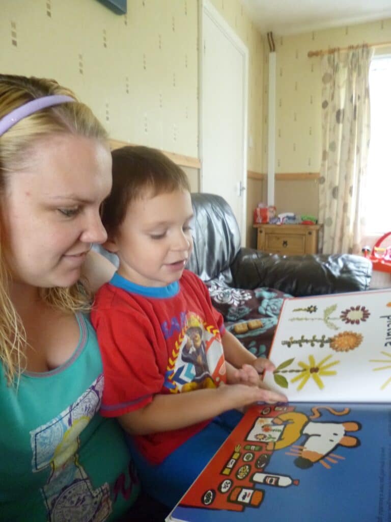 A woman and a young boy sit closely together on a sofa, happily reading a colourful Maisy craft book. The boy points at the pages filled with Maisy activities, while the cosy room features patterned curtains and toys in the background.