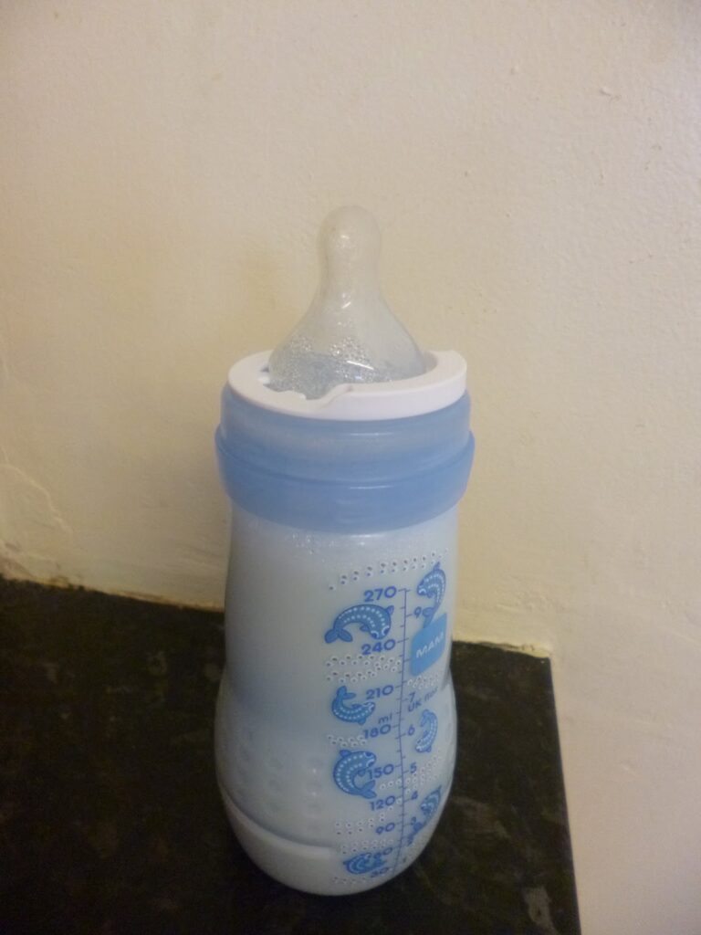 A baby bottle filled with milk, featuring a blue cap and teat, with measurement markings on the side, sits on a dark surface next to a Baby Nose Clear device against a light-coloured wall.