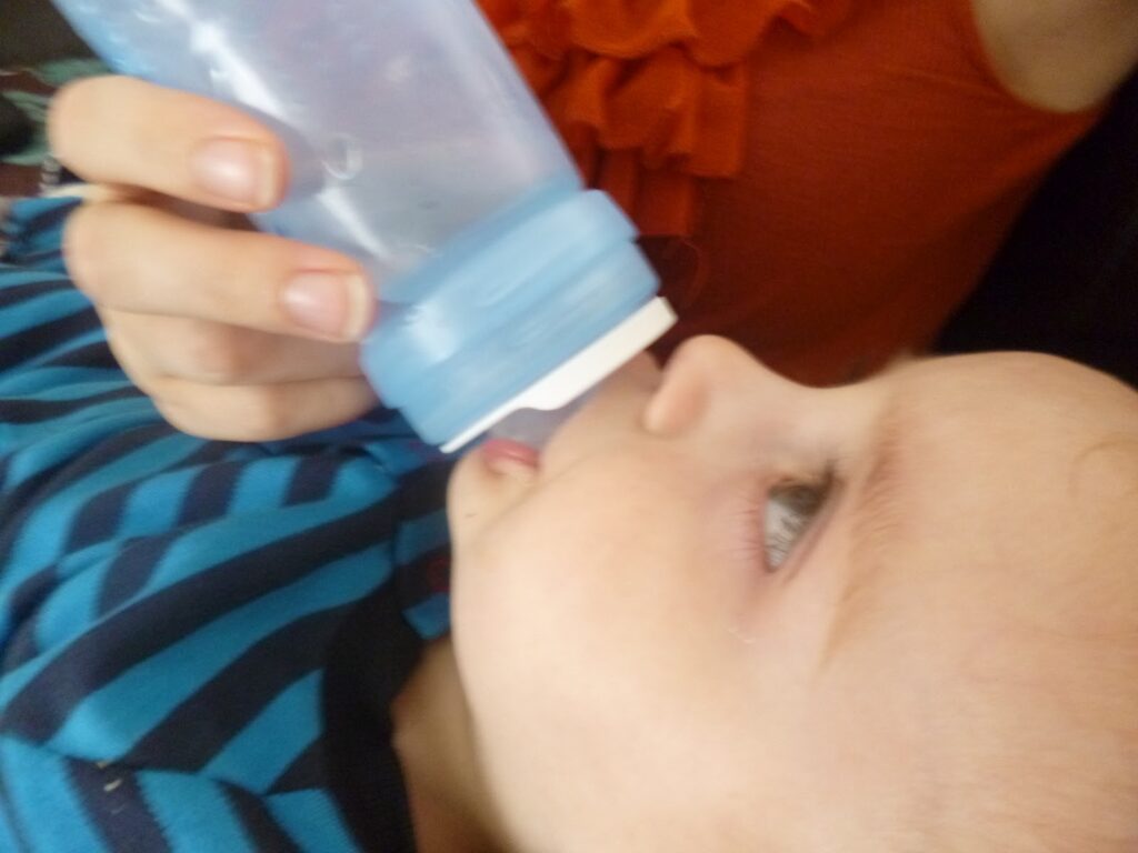 A close-up of a baby lying down and drinking from a bottle held by an adult in an orange top. The baby, wearing a blue and black striped shirt, looks comfortable thanks to recent Baby Nasal Relief.