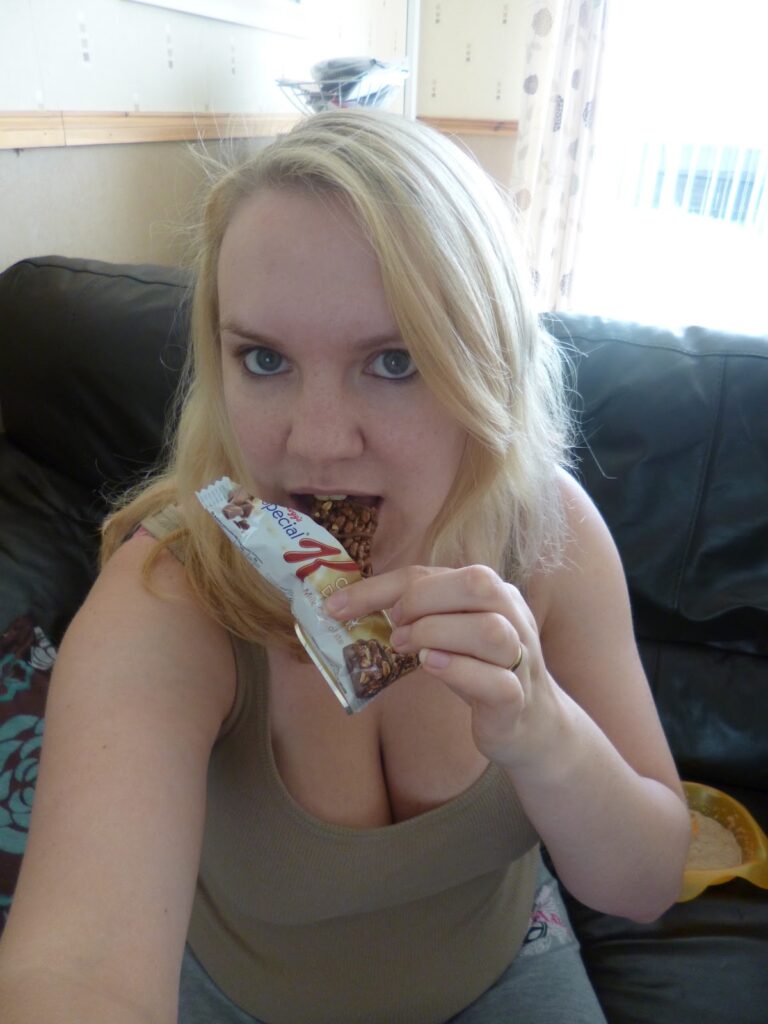 A woman with long blonde hair sits on a dark sofa, holding and eating a cereal bar whilst looking at the camera. She is wearing a beige vest top, enjoying a healthy snack as part of her post-pregnancy weight loss journey in natural indoor light.