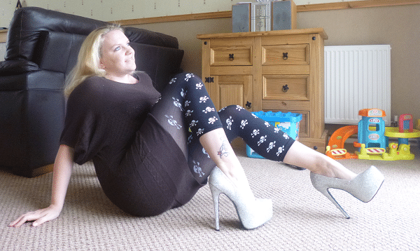 A woman with blonde hair, wearing a brown dress, floral leggings, and silver high heels, sits on a carpeted floor in a lounge with wooden furniture and children's toys in the background.