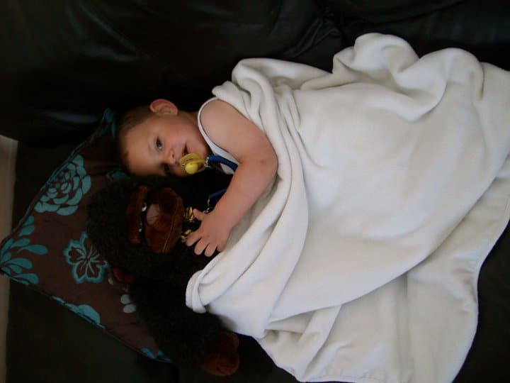 A young child lies on a sofa under a white blanket, holding a dummy and cuddling a stuffed black gorilla toy, as if ready to fight the monster under your bed, with their head resting on a blue and brown patterned cushion.