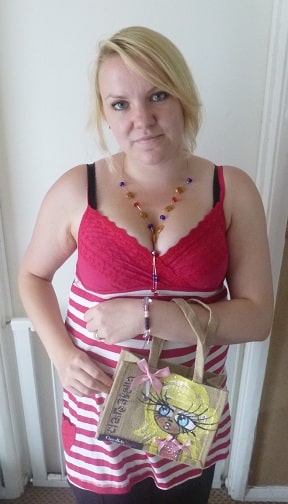 A woman with blonde hair in a red and white striped dress, red lace top, and beaded necklace holds a Polo Jute Bag with a cartoon face and "Glamour" on it. She stands indoors against a white wall.