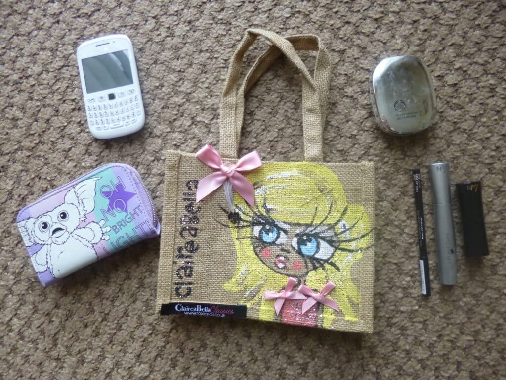 A small jute bag with a cartoon girl's face and pink bows sits on a carpet, surrounded by a white mobile, a purple Toxic Fox tin with an owl, a metallic compact, mascara, and lipstick.