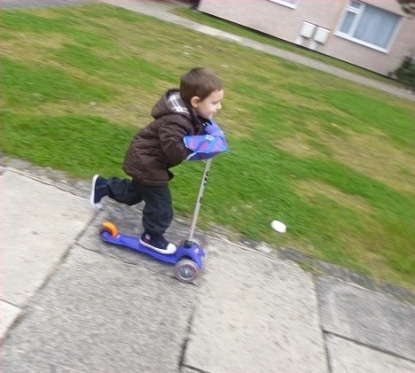 A young child wearing a brown jacket rides a blue and purple scooter on a quiet pavement for Silent Sunday, gliding past a grassy area in a peaceful residential neighbourhood.