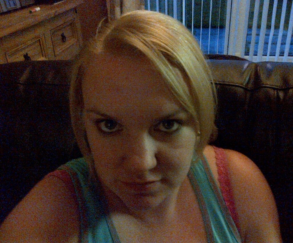 A woman with blonde hair and blue eyes, her locks shining as if nourished by macadamia oil, sits on a dark leather sofa indoors, looking directly at the camera. Sliding glass doors and a wooden cupboard are visible in the background.