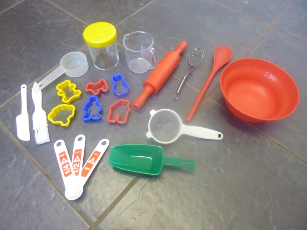A collection of toy baking tools from the Aldi Children's Baking Set on a tiled floor, including a mixing bowl, rolling pin, whisk, spoon, measuring spoons, biscuit cutters, sieve, and small containers.