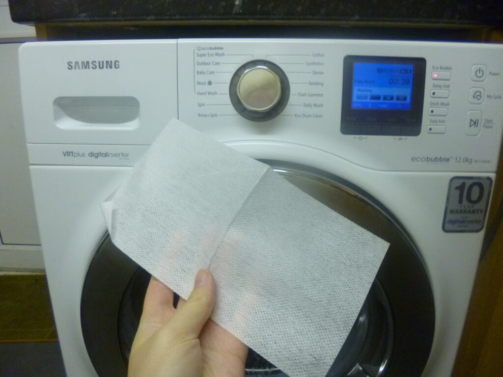 A hand holds a tumble dryer sheet in front of a Samsung front-loading washing machine with a digital display and control knob—perfect for use alongside stain remover or other cleaning products.