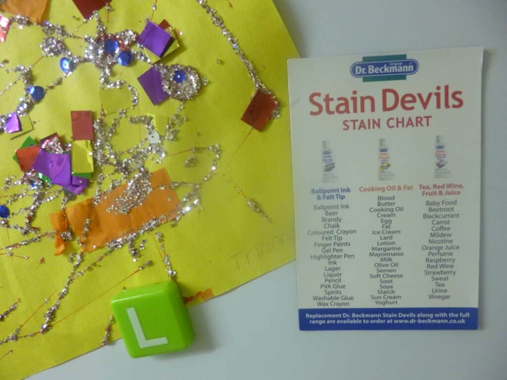A colourful craft with sequins, glitter, and confetti on yellow paper sits beside a Dr Beckmann Stain Devils stain chart and a green tile with the letter "L." The word "Franka" is faintly written, highlighting the need for a reliable stain remover.