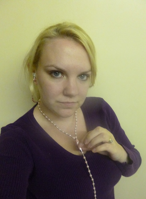 A woman with blonde hair wearing a purple top poses in front of a plain light-coloured wall, holding a Jewelfone Passion beaded necklace in her hand.
