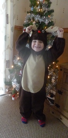 A young child smiles and poses in a festive Christmas Reindeer Costume in front of a decorated tree, holding up the costume’s antlers with both hands.