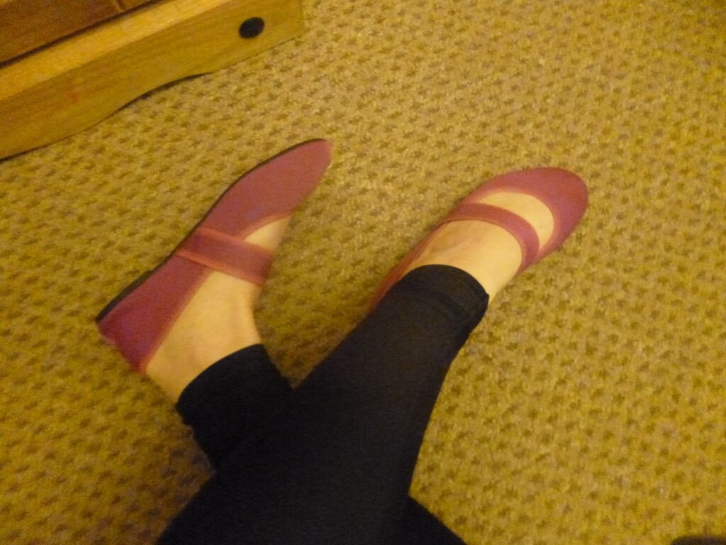 Person wearing black leggings and pink Mary Jane-style shoes sits on a beige carpeted floor near a wooden piece of furniture.