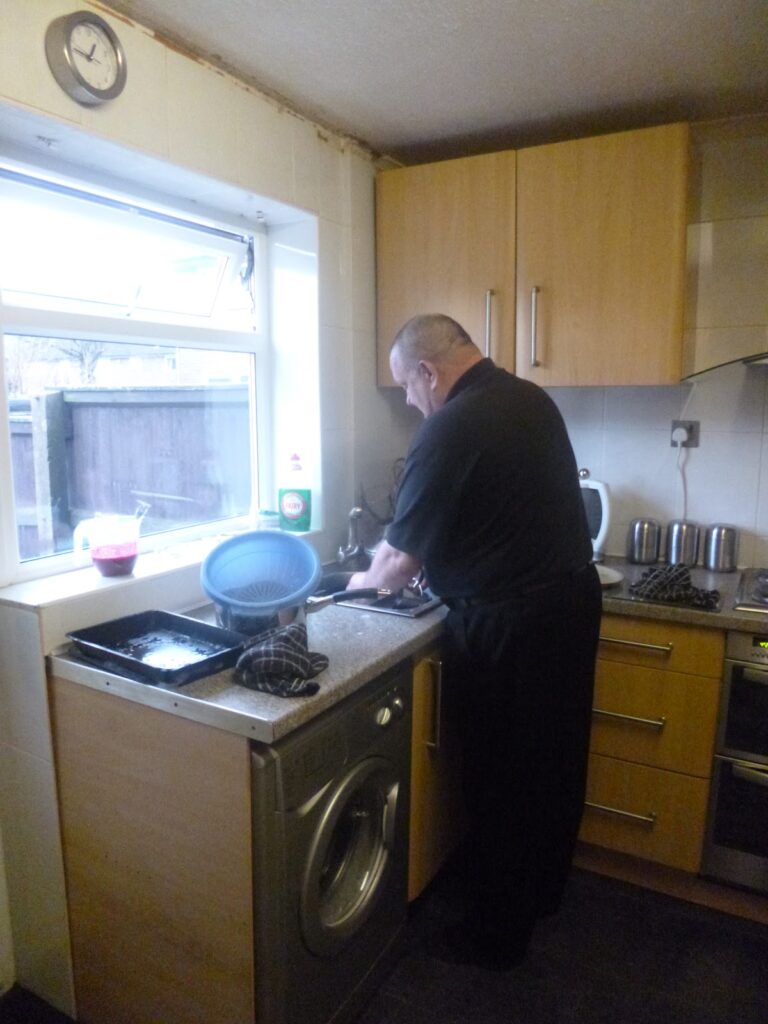 A man stands at a kitchen sink washing up beside a window on Christmas Day. There is a washing machine under the worktop, a draining rack with dishes on the worktop, and wooden cupboards and appliances in the background.