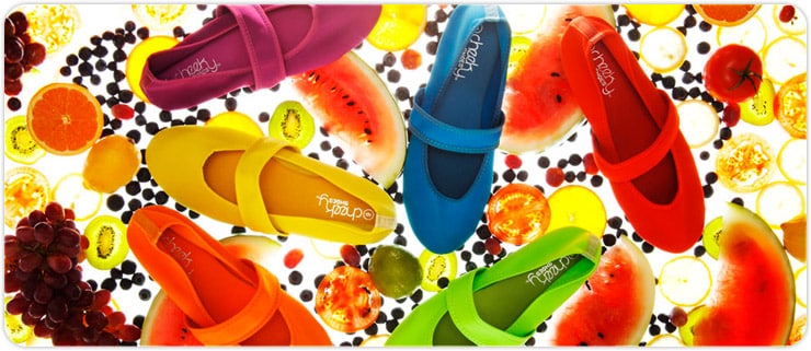 Six brightly coloured shoes (pink, yellow, orange, blue, green, and red) are arranged in a circle on a background filled with various fruits like watermelon, orange slices, grapes, lime, and tomatoes.