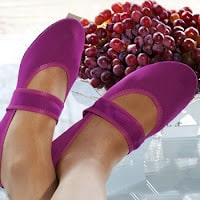 A person wearing bright purple Mary Jane shoes rests their feet on a glass table, with a bunch of red grapes in the background.