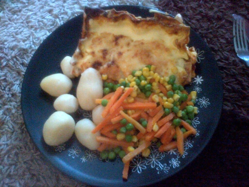A plate with a slice of lasagne—classic Italian food—several boiled potatoes, and a mix of carrots, peas, and sweetcorn on a black plate with white floral patterns. A fork is visible in the background.