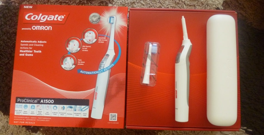 A Colgate ProClinical A1500 electric toothbrush set, featuring the toothbrush, a brush head, and a white travel case, is displayed next to the product box on a brown surface.