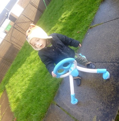 A young child wearing a tan animal-themed hat and dark jacket stands on a paved path, holding a blue and white Pewi walker, with green grass and a wooden fence in the background.