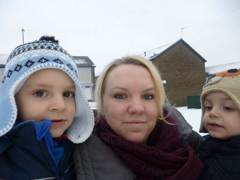 A woman with blonde hair and a maroon scarf poses outdoors with two young children in winter coats and hats, set against a snowy suburban backdrop with houses. It’s the perfect Let It Snow moment.