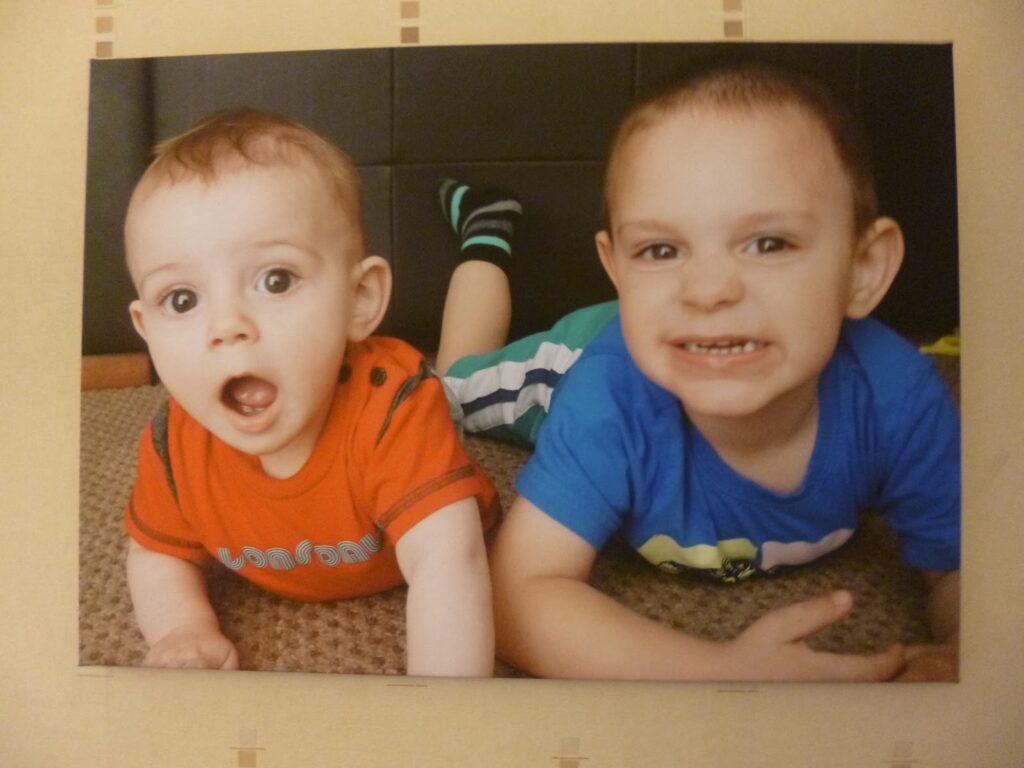 Two young children lie on the floor—one in a red shirt with a surprised expression, the other in blue smiling widely. Both look towards the camera, appearing happy and playful during their HelloCanvas giveaway photo moment.