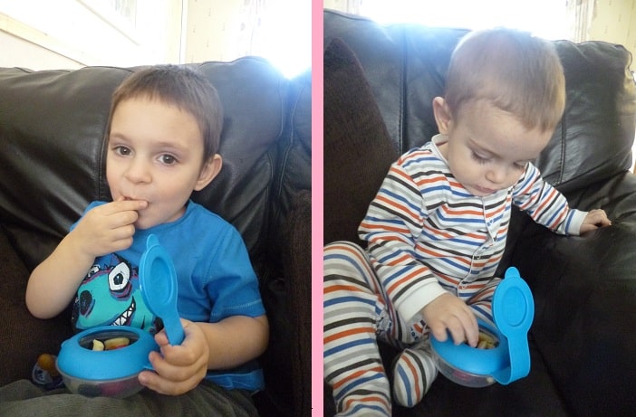 Split image: On the left, a young child in a blue shirt enjoys snacks from a MAM Snack Box on a brown sofa. On the right, a baby in striped pyjamas sits on the same sofa, reaching into a similar blue MAM Snack Box.