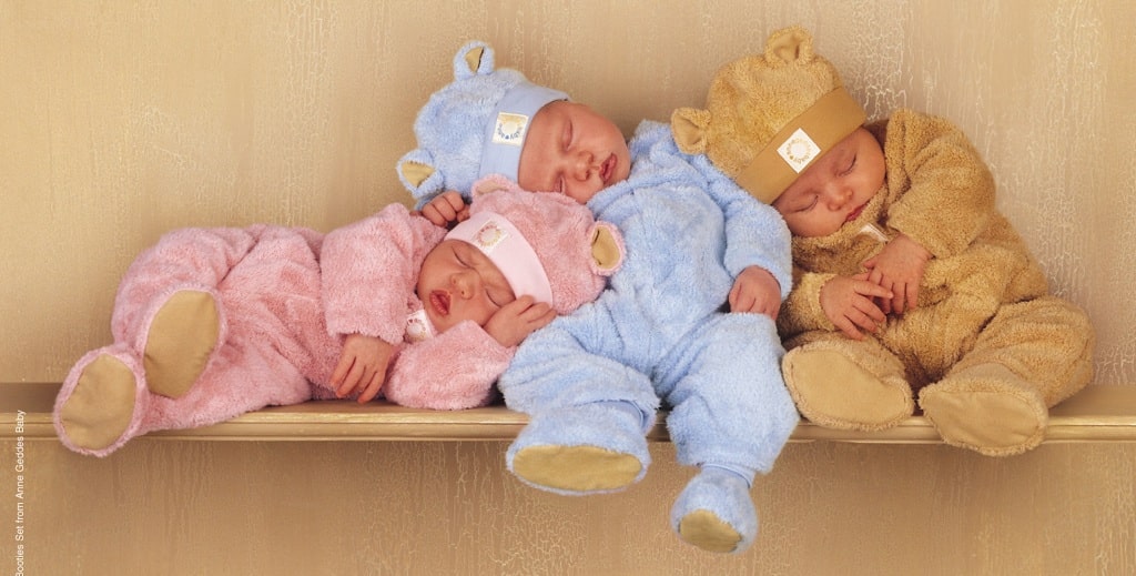Three sleeping babies dressed in fuzzy bear onesies—one in pink, one in blue, and one in brown—sit and lean against each other on a wooden shelf against a beige background, capturing the sweet parenting realities of peaceful moments.