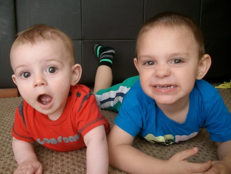 Two young children lie on a carpeted floor. The baby on the left in a red shirt looks surprised, whilst the older child in blue smiles widely at the camera, capturing a perfect moment of sibling joy.
