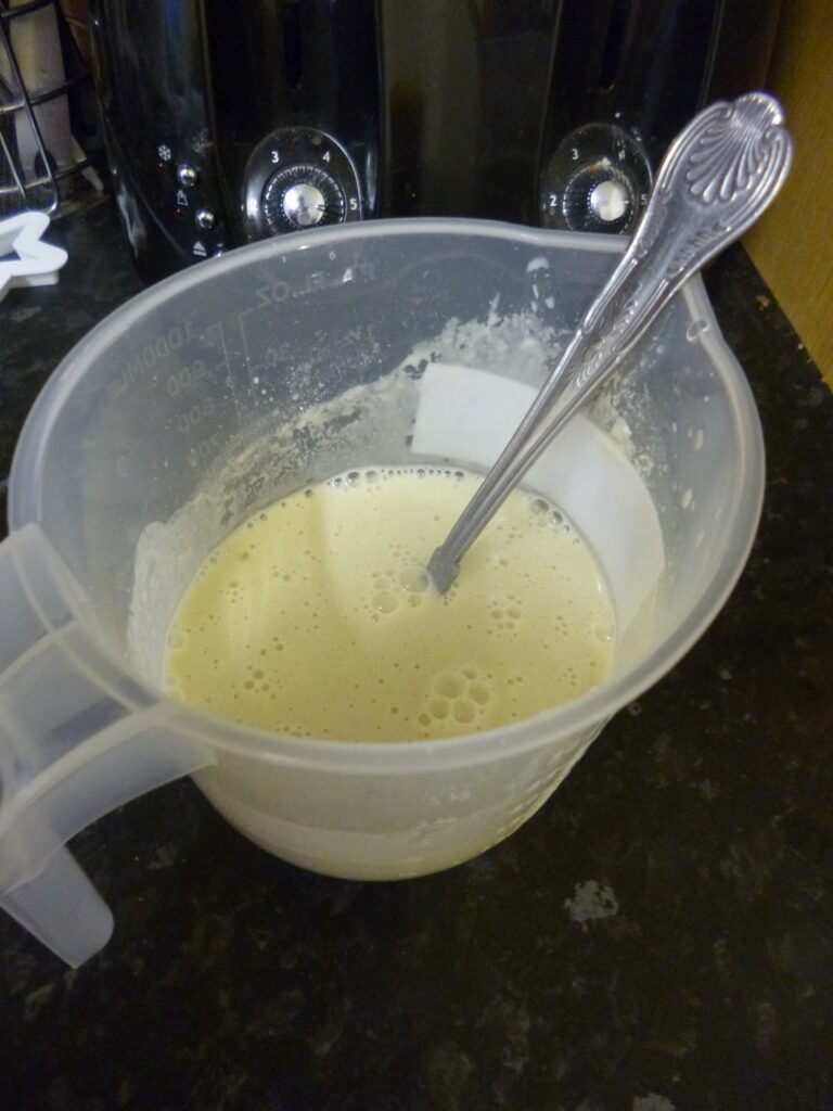 A plastic measuring jug containing a pale, frothy Flipping Stars batter with a metal spoon inside, placed on a dark kitchen worktop.