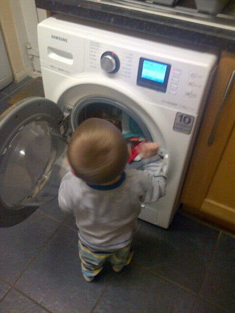 A small child stands in front of an open front-loading washing machine, holding clothes and appearing to help with the laundry—showing that fun in the kitchen can extend to chores, too. The machine door is open and the child is seen from behind.