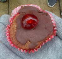 A hand in a grey glove holds a cupcake with chocolate icing and a red cherry on top, in a pink paper liner. Part of a shoe and pavement are visible in the background at a Red Nose Day charity event.
