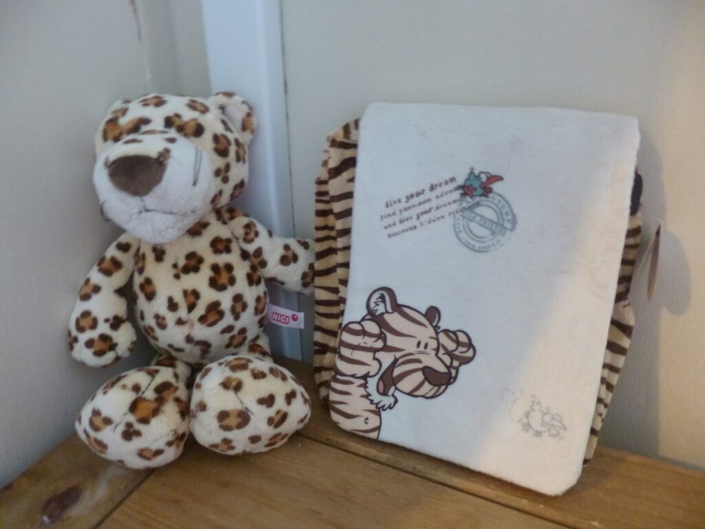 A plush leopard toy from NICI Wild Cats sits next to a cream-coloured notebook with a cartoon tiger illustration and text on its cover, placed on a wooden surface against a light-coloured wall.