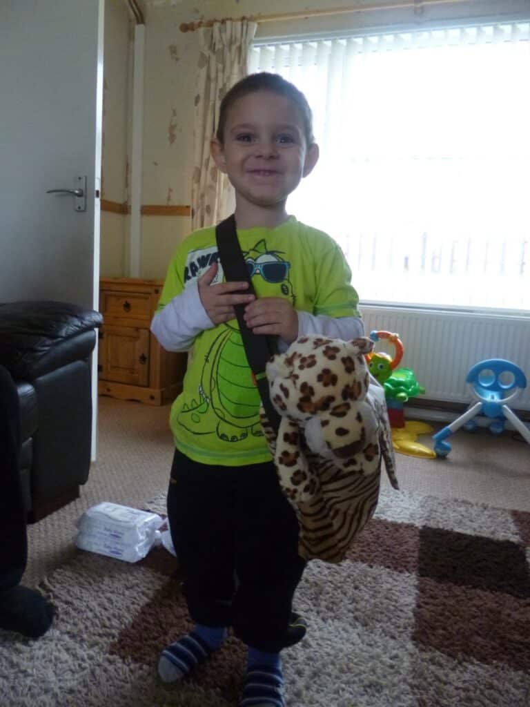A young child stands indoors, smiling in a bright green dinosaur shirt. The child holds a bag with a NICI Wild Cats stuffed leopard toy peeking out. Toys and furniture are visible in the background of the cosy room.