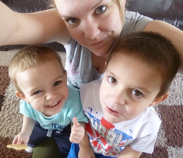 A woman takes a selfie with two young boys, all smiling and sitting close together on a patterned carpet in their newly completed house extension. One child is holding a small biscuit and giving a thumbs-up.
